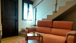 Una sala de estar con un sofá, una mesita, un tapete rosa, piso de madera, una puerta oscura y una escalera.