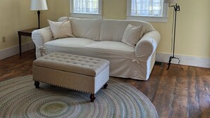A living room with a beige sofa, a matching ottoman, a patterned rug, two windows with sheer curtains, a floor lamp, and a framed picture on the wall. - Historic Cape in quaint village of Jefferson Maine (Jefferson)