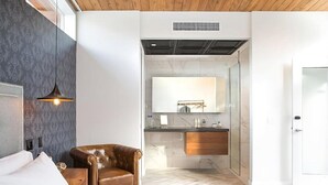 A modern hotel room with a leather armchair, a large bed, a bathroom with a glass shower, and a wooden ceiling. - New 3BR Apartment in Riga, Latvia (Riga)