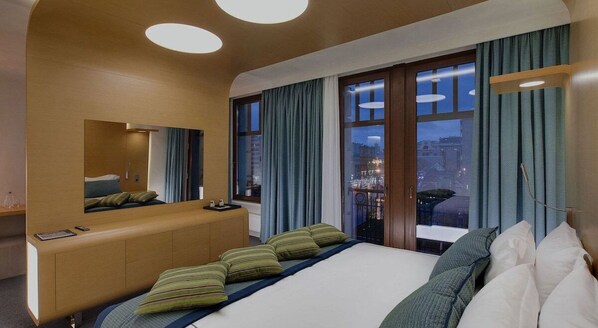 A modern hotel room with a large bed, a wooden headboard, a mirror, and a view of the city at night.