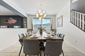 Een eetkamer met een grote tafel voor zes personen, omringd door stoelen, een kroonluchter en kunstwerken aan de muren.