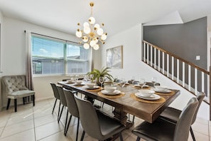 A dining area with a long wooden table set for six, surrounded by chairs, a chandelier, and a staircase in the background. - Sleeps 22•Pvt pool & spa•Game room (Kissimmee)