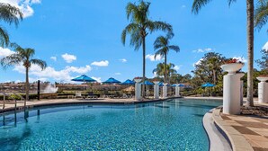 Ein Swimmingpool mit Liegestühlen und Sonnenschirmen, umgeben von Palmen und blauem Himmel.