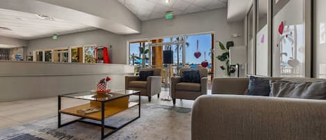 A modern lobby with a reception desk, seating area, and large windows offering a view of palm trees.