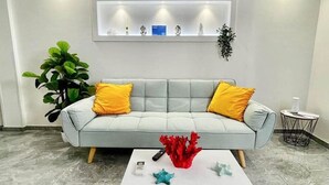 Un salon moderne avec un canapé gris, des coussins jaunes, une table basse blanche sur laquelle sont posés des livres, et une étagère murale agrémentée d’objets décoratifs.