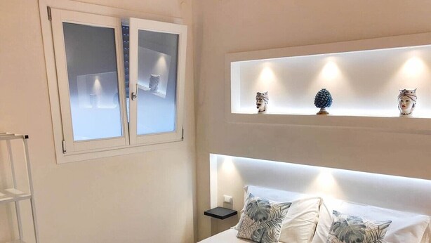 Un lit bien fait, avec du linge de lit blanc et deux serviettes, une table de chevet avec une lampe et une étagère ornée d’objets décoratifs.