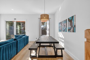 A modern living room with a teal sofa, wooden floor, a central table with benches, a hanging light fixture, and abstract wall art. - Chic Townhome w/ Pool & Game Room | Near Downtown! (St. Augustine)