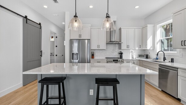 Une cuisine moderne dotée d’un grand îlot central, d’appareils électroménagers en acier inoxydable et de luminaires suspendus.