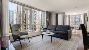 A modern living room with a grey sofa, a round coffee table, and a grey armchair. Large windows offer a view of high-rise buildings.