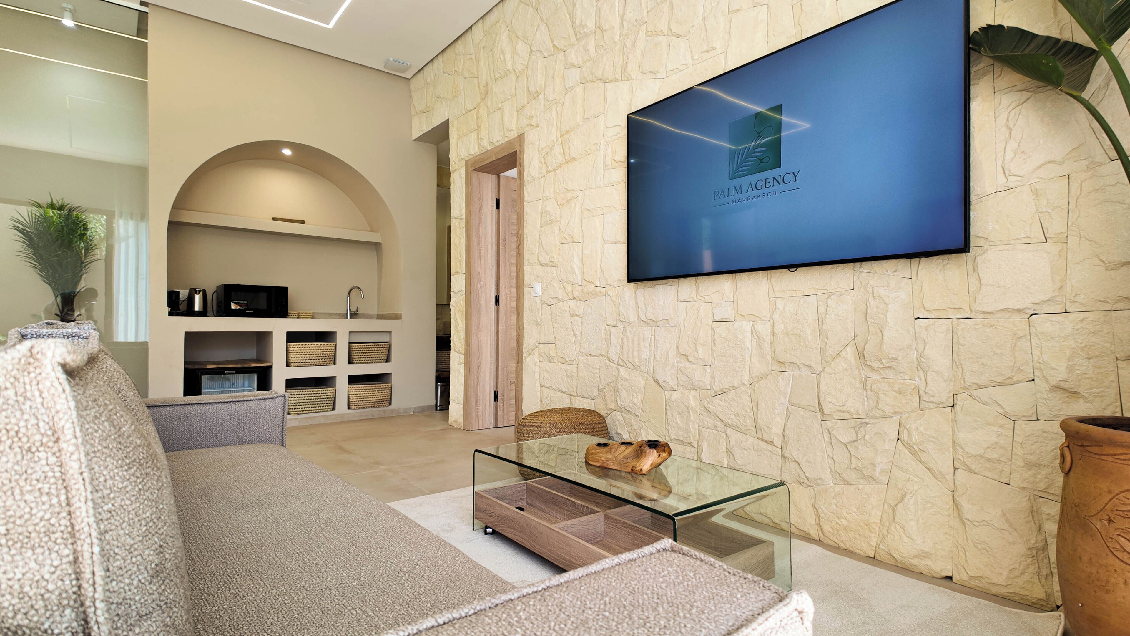 A modern lobby with a stone wall, a flat-screen TV displaying 'Palm Agency Marrakech', a glass coffee table, and a cozy seating area.