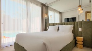 Una cama grande con cabecera verde, ropa de cama blanca y vistas a una piscina a través de cortinas translúcidas.