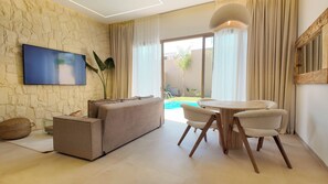 Una sala de estar moderna con pared de piedra, un televisor de pantalla plana, un sofá, una mesa de comedor con sillas y vistas a una piscina y jardín.