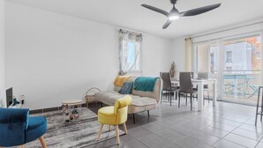 A modern living room with a sofa, yellow armchair, and blue chair. There is a dining area with a table and chairs. A ceiling fan is visible. - Appartement calme et lumineux, terrasse et P privé (Balma)