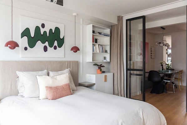 Une chambre moderne avec un grand lit, une bibliothèque et un coin repas visible à travers une porte ouverte.
