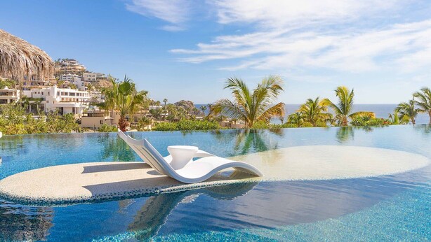 Ein Hotelpool mit klarem, blauem Wasser, weißen Liegestühlen und Blick auf das Meer und Gebäude im Hintergrund.