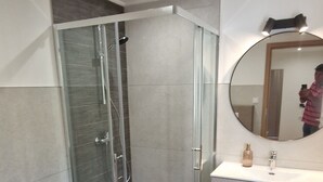 Un bany modern amb dutxa envidriada, un lavabo blanc i un mirall rodó.