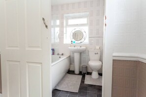 Ein Badezimmer mit einem weißen Waschbecken, einer Toilette und einer Badewanne. Über dem Waschbecken befindet sich ein Fenster, und eine Tür führt in einen anderen Raum.
