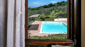 A view through a window of a swimming pool, greenery, and a book on a table. - Villa with pool surrounded by an olive grove (Assisi)