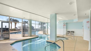 An indoor swimming pool with clear glass walls, a tiled floor, and a poolside area with chairs and a sunshade. - The Dunes 100 (Gulf Shores)