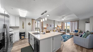 A modern kitchen with white cabinets, a central island, and stainless steel appliances. There is a dining area with a patterned rug and a sofa. - The Dunes 100 (Gulf Shores)