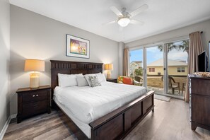 A bedroom with a large bed, two bedside tables with lamps, a ceiling fan, a window with a view of houses, and a sliding glass door. - The Dunes 100 (Gulf Shores)