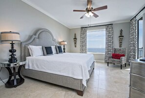 A bedroom with a large bed, two bedside lamps, a ceiling fan, a view of the ocean, a zebra-patterned chair, and a dresser. - Indigo West 502 (Pensacola)