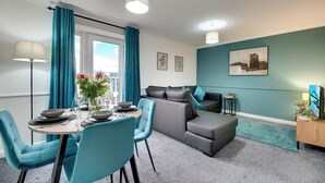 Uma sala de estar moderna com sofá cinza, cortinas verde-água e um espaço de jantar com cadeiras verde-água e uma mesa posta para dois.