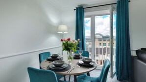 Uma área de jantar com mesa redonda posta para quatro pessoas, cadeiras verde-água, um vaso com flores e vista para um horizonte urbano através de uma janela grande com cortinas verde-água.