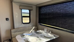 A dining area in a camper with a table set for two, a window with blinds, and a small trash can. - Cozy 2-bedroom brand new trailer in quaint Whitewater with all amenities. (Whitewater)