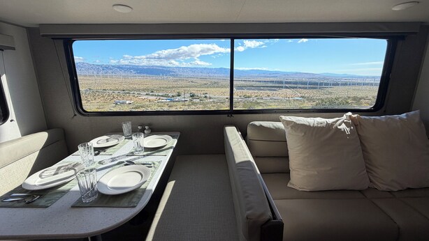 A travel trailer interior with a dining area, a sofa, and a large window offering a view of a desert landscape with mountains. - Cozy 2-bedroom brand new trailer in quaint Whitewater with all amenities. (Whitewater)