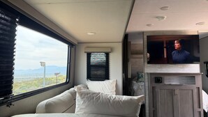 A modern RV interior with a large window, a flat-screen TV, and a cozy seating area. - Cozy 2-bedroom brand new trailer in quaint Whitewater with all amenities. (Whitewater)