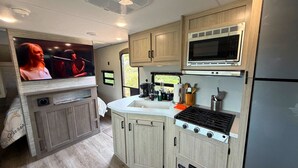 A compact RV kitchen with a microwave, stove, sink, and a TV mounted on the wall. - Cozy 2-bedroom brand new trailer in quaint Whitewater with all amenities. (Whitewater)