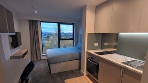 Ein modernes Hotelzimmer mit Bett, Schreibtisch, einer Küchenzeile und einem großen Fenster mit Blick auf die Stadt.