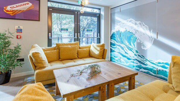 Eine moderne Hotelhalle mit einer großen Wandmalerei im Wellen-Design, einer Couch, einem Holztisch und einem Essbereich mit Stuhl und Tisch.