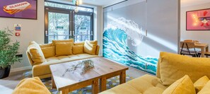 A modern hotel lobby with a large wave mural, a sofa, a wooden coffee table, and a dining area with a chair and table.