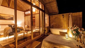 Ein gemütliches Blockhaus mit einer großen Glasschiebetür, die zu einem Schlafzimmer, einer Holzterrasse, einer Badewanne und einem Essbereich mit Tisch für zwei führt.