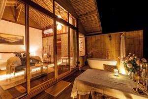 Un chalet confortable doté d’une grande porte-fenêtre coulissante donnant sur une chambre, d’une terrasse en bois, d’une baignoire et d’un coin repas avec une table dressée pour deux personnes.