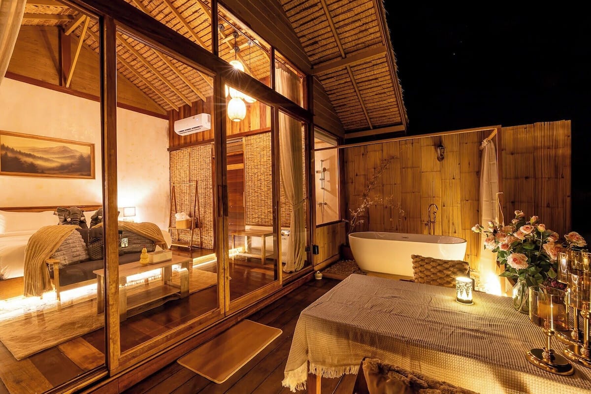 Una cabaña acogedora con un amplio ventanal corredera que da a un dormitorio, una terraza de madera, una bañera y un comedor con una mesa para dos.
