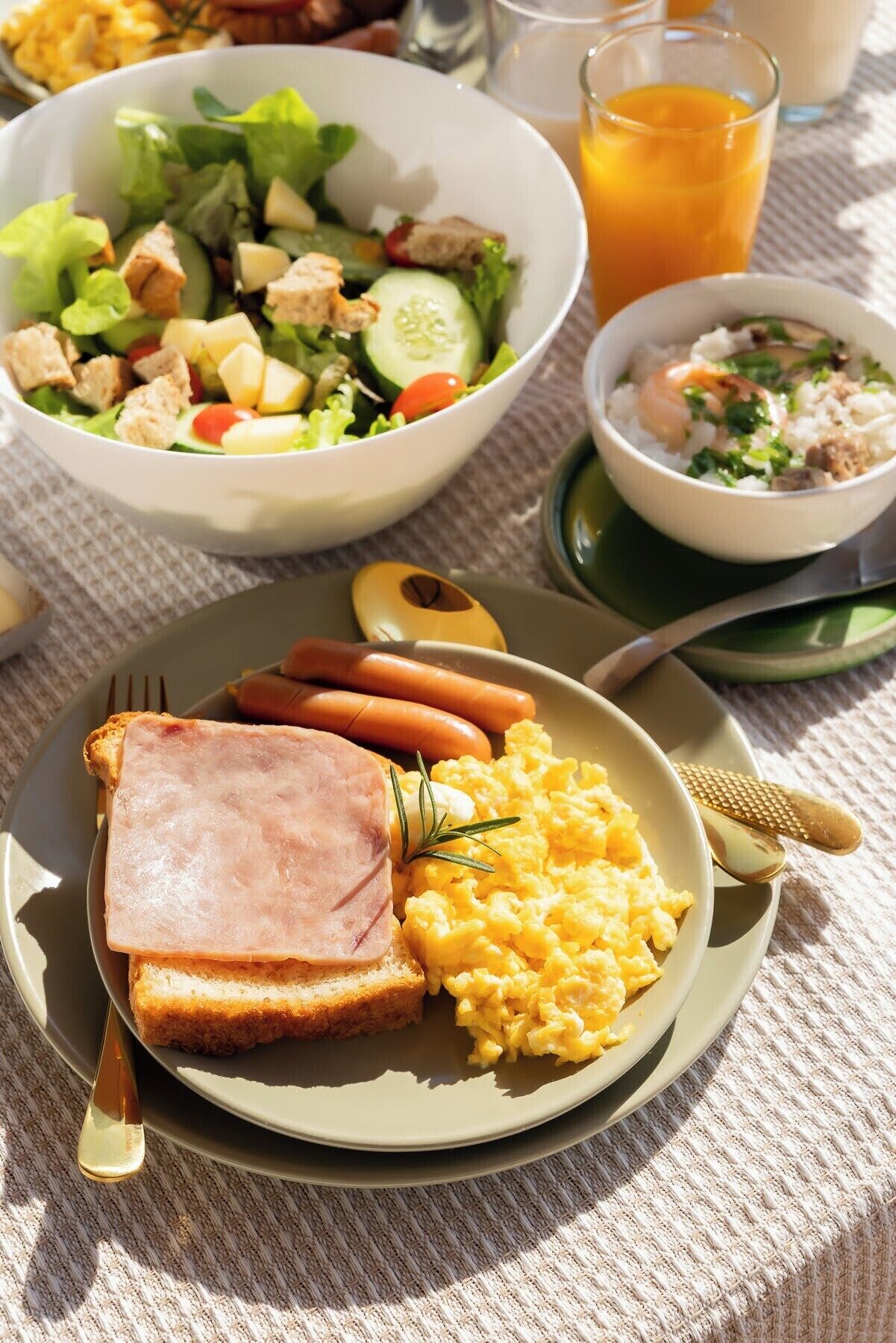 A breakfast plate with ham, scrambled eggs, and toast, a bowl of salad, and a glass of orange juice.