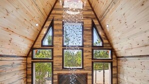 L’intérieur d’un chalet en bois, avec une cheminée centrale, de nombreuses fenêtres et un luminaire au plafond.