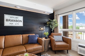 A modern living room with a black wall, a city limit sign, a leather sofa, a brown armchair, a side table with a lamp, and a large window with a view. - The Landing Loft! Walk to Downtown, Pools, Fishing, Gym, Scenic Train Station.. (Branson)