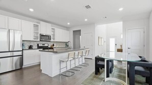 Una cocina moderna con armarios blancos, una isla central con taburetes y una mesa de comedor de vidrio, además de luces empotradas en el techo.