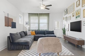 A modern living room with a sectional sofa, a small armchair, a wooden bench, and a large TV. - Porch House Retreat (Travis County)
