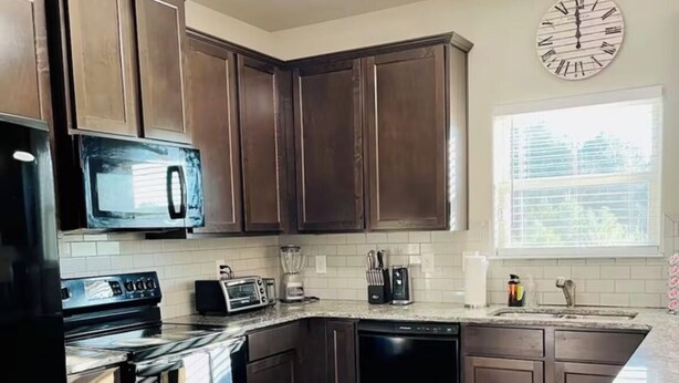 A modern kitchen with dark wood cabinets, a black refrigerator, a microwave, a stove, a dishwasher, and a sink. There is a clock on the wall and a window providing natural light. - Cozy 4BR + Fast WiFi + Self Check-In + Near I-75 (McDonough)