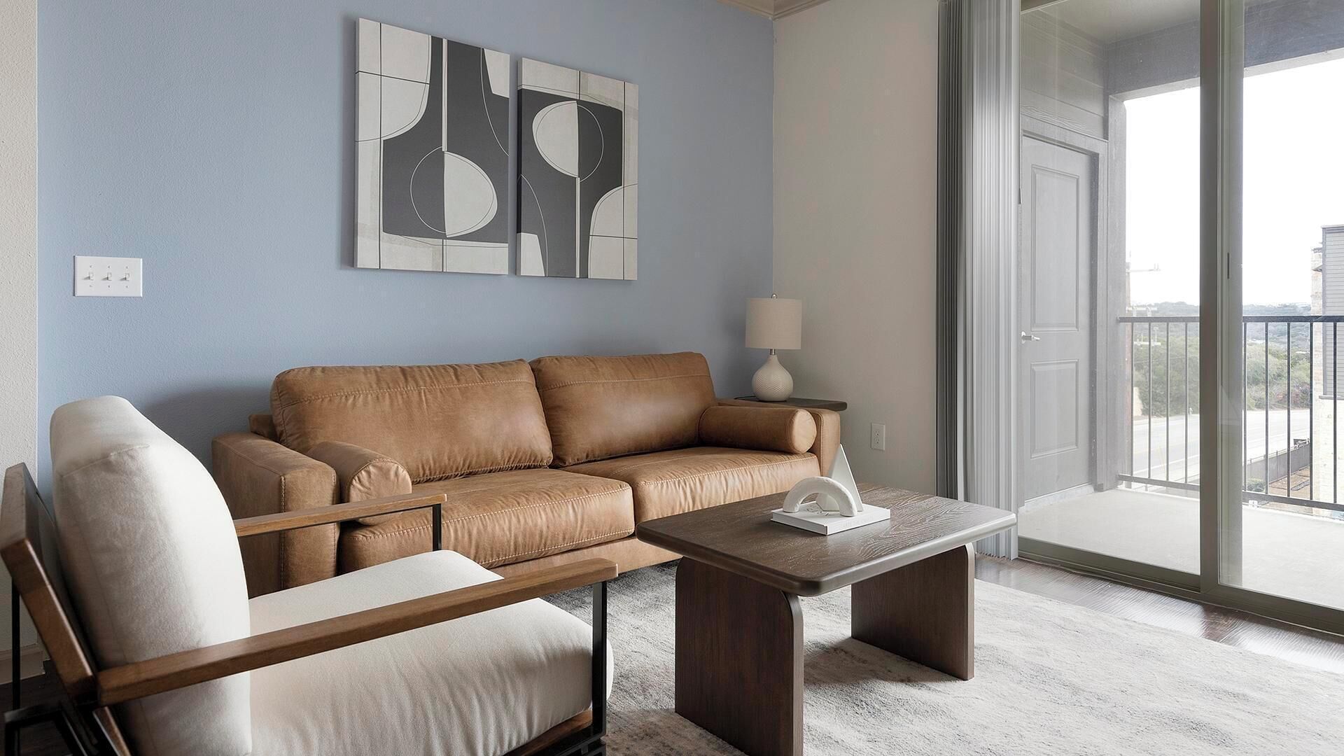 Un salon avec un canapé en cuir marron, une table basse en bois et un fauteuil blanc. Un tableau abstrait moderne est accroché au mur, et une fenêtre offre une vue sur l’extérieur.