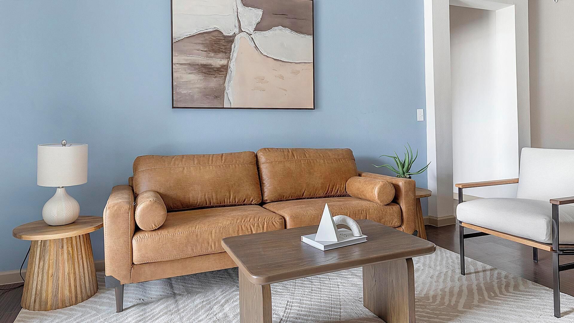 Un salon avec un canapé en cuir marron, une table basse en bois, un fauteuil blanc, une petite table d’appoint avec une lampe et une plante en pot.