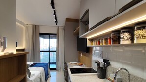 Ein modernes Hotelzimmer mit Küchenzeile, einem Bett mit blauer Kopfteil, einem großen Fenster mit Vorhängen und einem an der Wand montierten Fernseher.
