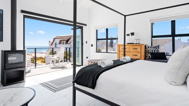A modern bedroom with a large bed, a TV, a wooden dresser, and a balcony with a view of the sea. - C’est La Vie Beach Villa (Saint Helena Bay)