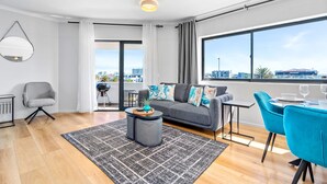 Ein modernes Wohnzimmer mit grauem Sofa, einem runden Couchtisch, einem Esstisch mit blauen Stühlen und einem großen Fenster mit Blick auf die Stadt.