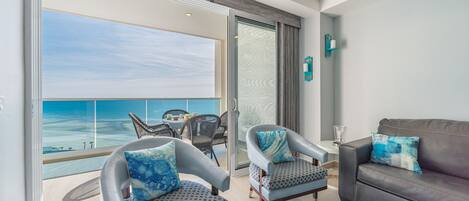 A balcony with a view of the ocean, two armchairs with patterned cushions, a grey sofa with matching cushions, and a small table with a glass.